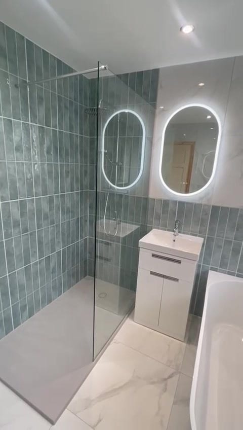 Shower room with glass walk-in screen, white vanity basin unit and oval LED mirror on sage tiled wall